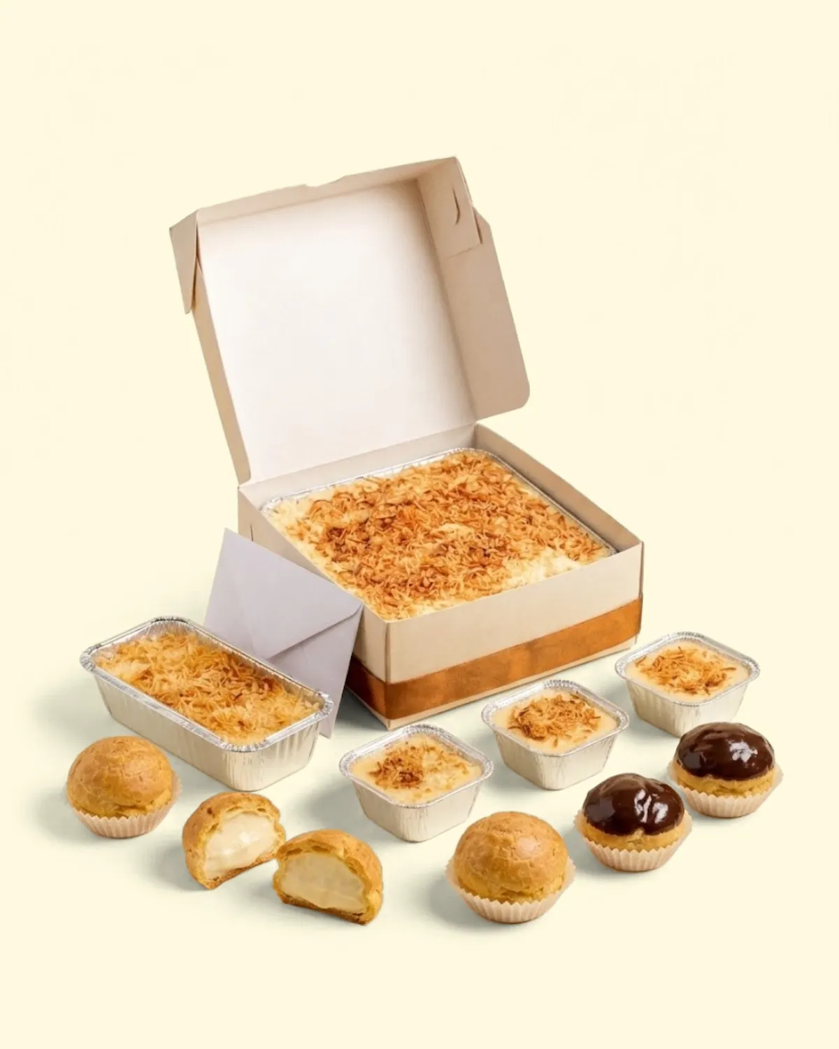 Box kado terbuka berisi klappertaart dengan pita dan amplop, dikelilingi klappertaart 500 g, klappertaart cup, soes original, dan soes cokelat